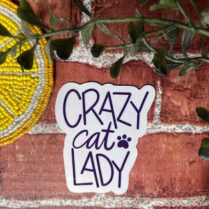 May include: White sticker with the text "CRAZY cat LADY" in purple lettering with a paw print next to the word "cat".