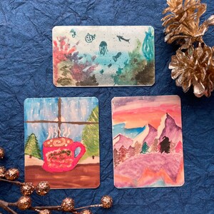 May include: Three rectangular watercolor paintings. One depicts an underwater scene with coral and marine life. Another shows a mug of hot cocoa by a snowy window. The third features a mountain landscape.