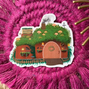 May include: A whimsical illustration of a small, brown house with a green roof covered in mushrooms. The house has a round door with a wreath and a small window with a light inside. The house is surrounded by green grass and a wooden fence.