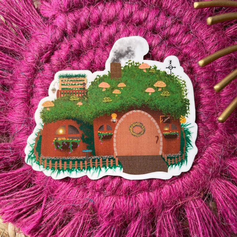 May include: A whimsical illustration of a small, brown house with a green roof covered in mushrooms. The house has a round door with a wreath and a small window with a light inside. The house is surrounded by green grass and a wooden fence.
