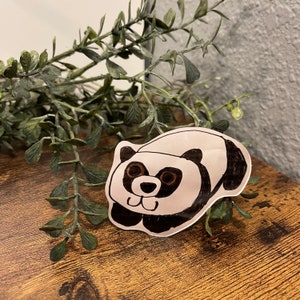 May include: A hand-drawn panda sticker with black and brown details. The panda is lying down with its paws extended. The sticker is on a wooden surface with green foliage in the background.