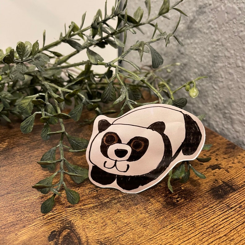 May include: A hand-drawn panda sticker with black and brown details. The panda is lying down with its paws extended. The sticker is on a wooden surface with green foliage in the background.