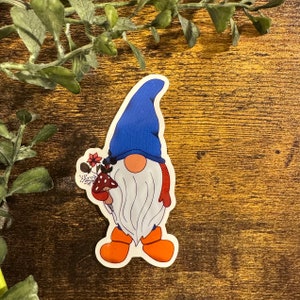 May include: A blue and white gnome with a red hat and orange boots holding a mushroom and flowers.