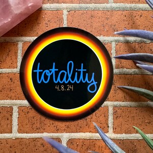 May include: A round sticker with the word "totality" in blue script, surrounded by a yellow and orange ring against a black background. The date "4.8.24" is below the word. The sticker is on a brick wall.