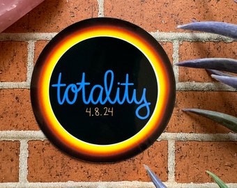 May include: A round sticker with the word "totality" in blue script, surrounded by a yellow and orange ring against a black background. The date "4.8.24" is below the word. The sticker is on a brick wall.