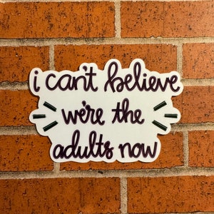 May include: White sticker with black text that reads "i can't believe we're the adults now". The sticker is on a brick wall.
