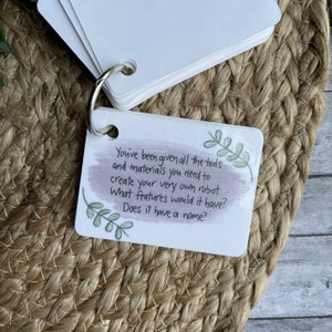 May include: A white card with green leaves and a watercolor background. The card has the text "You've been given all the tools and materials you need to create your very own robot. What features would it have? Does it have a name?"