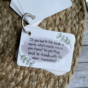 May include: A white card with a watercolor background and green leaves drawn on it. The card has the text "If you had to live inside a movie, which movie would you choose? Do you think you'd be friends with the main characters?"