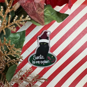 May include: A black cat wearing a Santa hat and sitting on a green and white oval-shaped ornament. The ornament has the text "Santa, let me explain".