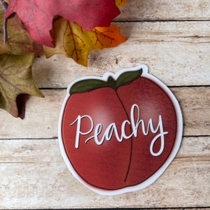 Pode incluir: Um adesivo em forma de pêssego com contorno branco e a palavra "Peachy" escrita em letras cursivas brancas.