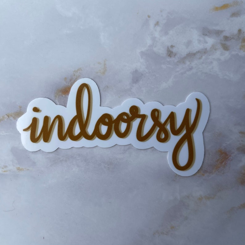 Op de afbeelding: Een gouden sticker met het woord "indoorsy" in een cursief schrift.