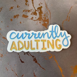 May include: A white sticker with the text "currently adulting" in blue and yellow lettering. The sticker has a white outline.