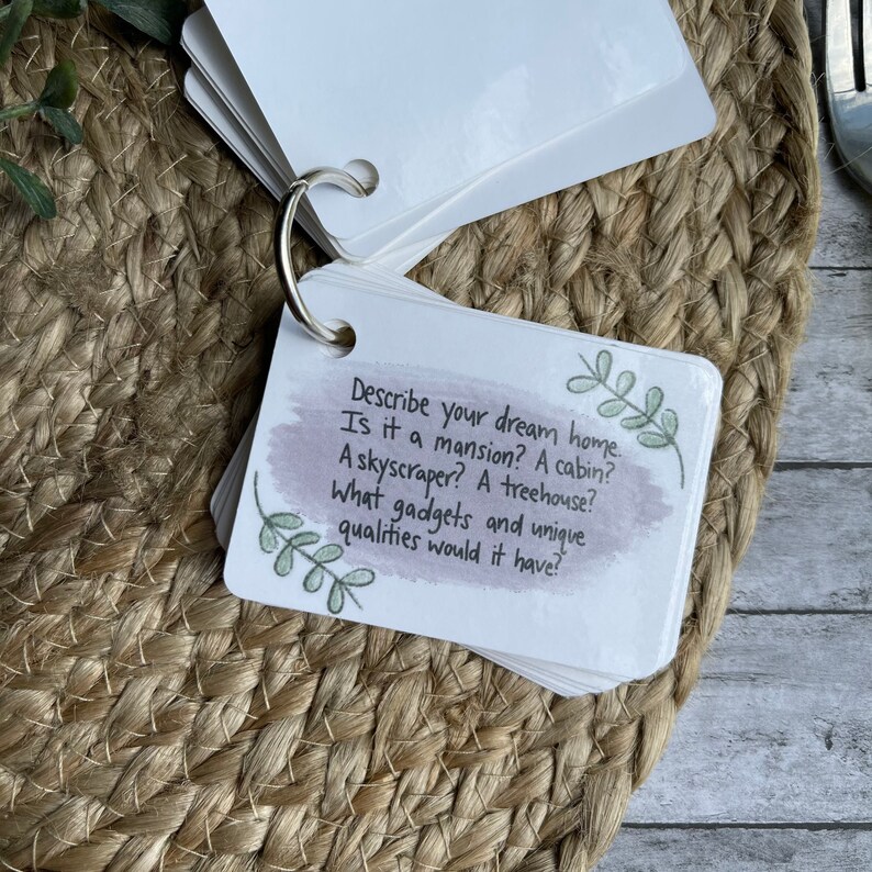 May include: A set of white cards with a ring binder. The cards have a watercolor background and a handwritten question: "Describe your dream home. Is it a mansion? A cabin? A skyscraper? A treehouse? What gadgets and unique qualities would it have?"