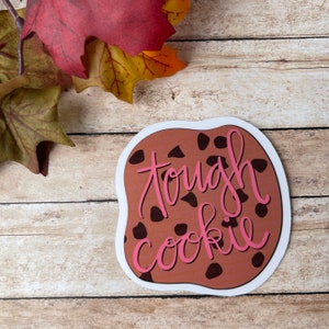 May include: A brown cookie sticker with the text "tough cookie" written in pink. The cookie has chocolate chips.