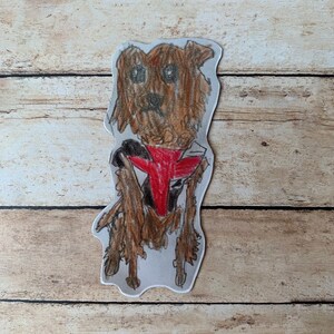 May include: A hand-drawn illustration of a dog with brown fur, wearing a red and black vest. The dog is drawn with a pencil and has a friendly expression. The artwork is cut out and placed on a wooden background.
