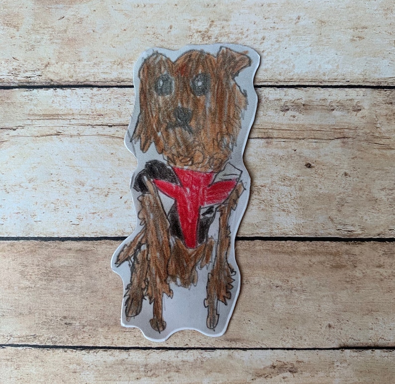 May include: A hand-drawn illustration of a dog with brown fur, wearing a red and black vest. The dog is drawn with a pencil and has a friendly expression. The artwork is cut out and placed on a wooden background.
