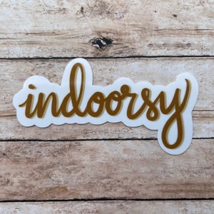 Op de afbeelding: Een gouden sticker met het woord "indoorsy" in een cursief schrift.