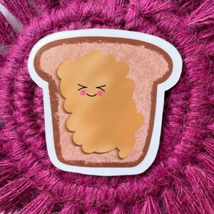 May include: A cartoon illustration of a slice of bread with peanut butter. The peanut butter has a smiling face with closed eyes and rosy cheeks. The bread is brown and the peanut butter is light brown.