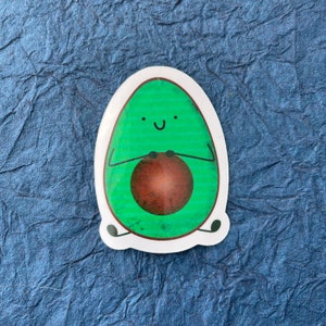 May include: A green avocado sticker with a smiling face and a brown pit. The avocado is holding its pit in its arms.