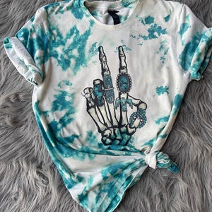 May include: White t-shirt with a turquoise tie-dye pattern. The shirt features a graphic of a skeleton hand making a peace sign, adorned with turquoise rings. The shirt is knotted at the bottom.