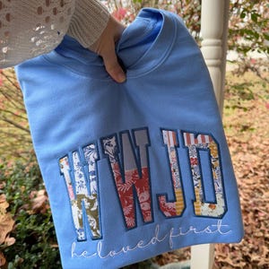 May include: A light blue sweatshirt with the embroidered text "WWJD" in a patchwork design of floral and patterned fabrics. The words "beloved first" are embroidered below in a cursive font.