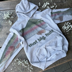 May include: A light gray hoodie featuring a large watercolor-style rainbow trout design on the front and sleeves. The text "that big, huh?" is printed below the fish. The hoodie has a hood and a front pocket.