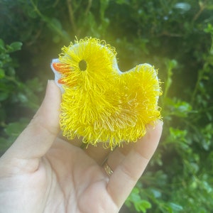 May include: A yellow fuzzy duckling with a red and orange beak and black eyes. The duckling is made of fabric and has a fluffy texture.