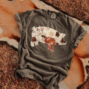 May include: Dark grey t-shirt featuring a patchwork pig design. The pig is composed of floral, sunburst, and cowhide patterns. The shirt has rolled-up sleeves and a crew neck. The background is a brown and tan animal hide.