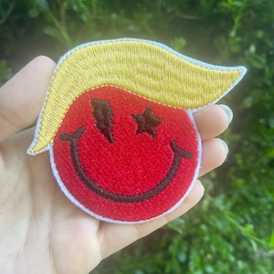 May include: A red embroidered smiley face with black eyes and a black outline. The smiley face has two stars for eyes and a yellow embroidered hairpiece on top.
