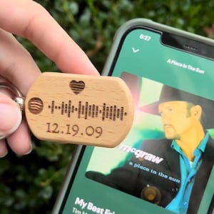 May include: A wooden keychain with a Spotify code, a heart, and the date 12.19.09. The keychain is held near a smartphone displaying a music app with the song "My Best Friend" by Tim McGraw.