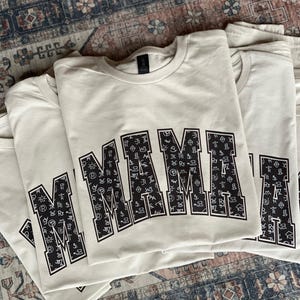 Stack of beige “MAMA” graphic sweatshirts featuring bold black varsity-style letters filled with white western cattle brand symbols. The design has a rustic ranch-inspired aesthetic, displayed on a vintage patterned rug background for a cozy, boho western vibe. Perfect gift for moms who love farmhouse or cowboy-inspired style.