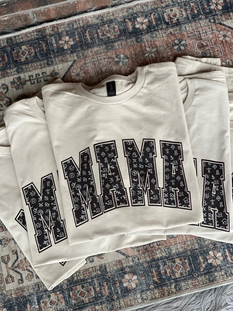 Stack of beige “MAMA” graphic sweatshirts featuring bold black varsity-style letters filled with white western cattle brand symbols. The design has a rustic ranch-inspired aesthetic, displayed on a vintage patterned rug background for a cozy, boho western vibe. Perfect gift for moms who love farmhouse or cowboy-inspired style.