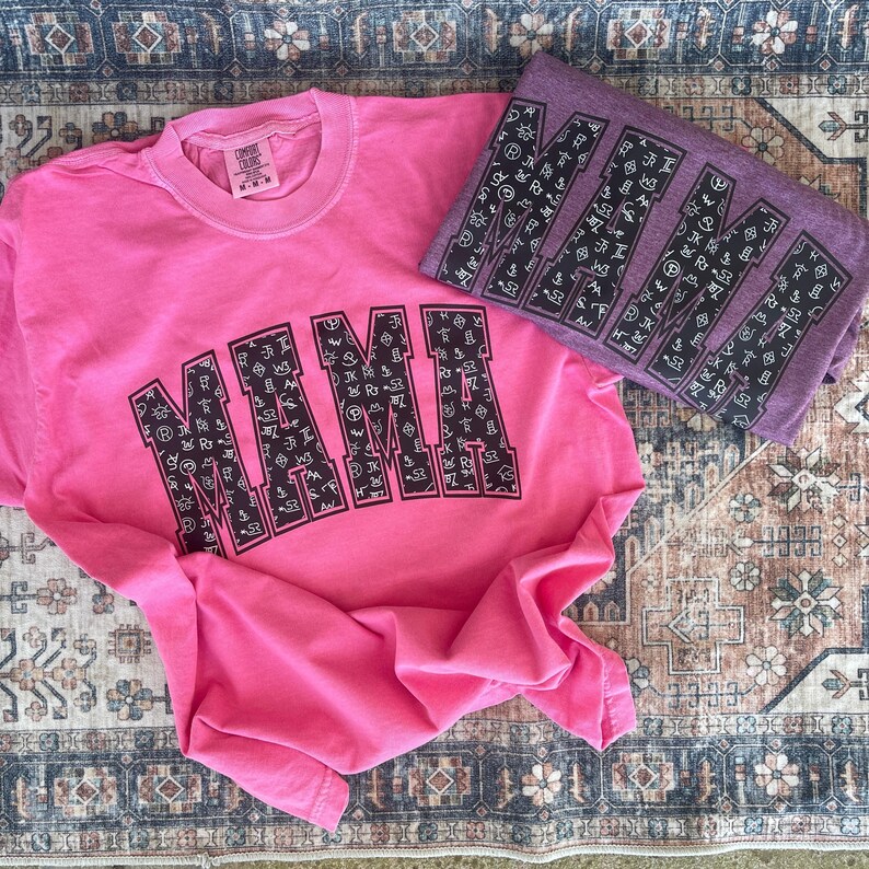 Bright pink and purple “MIMI” graphic sweatshirts featuring bold black varsity-style lettering filled with white western cattle brand symbols. The fun, rustic western design pops against the vibrant shirt colors, displayed on a vintage patterned rug background. Perfect cozy gift for grandmas who love a stylish boho cowgirl look.