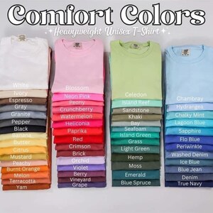 May include: Stacks of folded Comfort Colors heavyweight unisex t-shirts in various colors, including white, pink, green, and blue. Each stack displays a different color with a corresponding color name label. The image features the text "Comfort Colors".