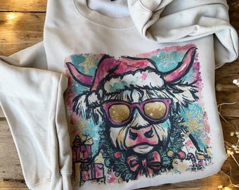 May include: Beige sweatshirt featuring a whimsical graphic of a Highland cow wearing a Santa hat and sunglasses. The cow is surrounded by snowflakes and gift boxes, with a red bow. A cozy, festive design.