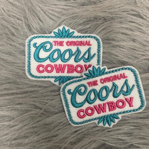 May include: Two rectangular embroidered patches with white fabric backgrounds. Each patch features the text THE ORIGINAL Coors in teal script and COWBOY in pink block letters, framed by a teal border with decorative teal floral accents.