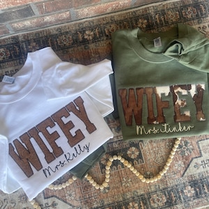 May include: Two white t-shirts with the word "WIFEY" in brown faux leather lettering. The shirt on the left has the text "Mrs. Kelly" underneath the word "WIFEY". The shirt on the right has the text "Mrs. Tinker" underneath the word "WIFEY". The word "WIFEY" is decorated with a cow print pattern.