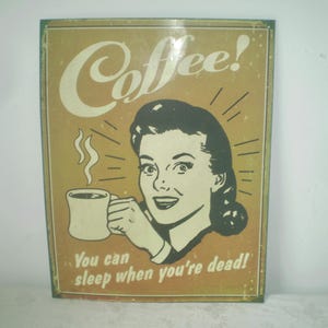 May include: Vintage-style metal sign with a retro illustration of a woman holding a coffee mug. The sign features the word "Coffee!" and the phrase "You can sleep when you're dead!" in white text on a tan background.