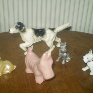 May include: A collection of five dog figurines including a large white and black spotted dog, a small metallic gold dog, a solid pink dog, a small grey textured dog, and a white dog with a red collar, all arranged together as a group.