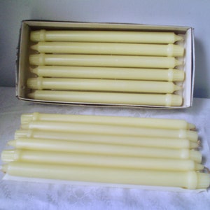Peut inclure: Plusieurs bougies coniques de couleur ivoire, certaines dans une boîte en carton, d'autres posées sur une surface blanche. Les bougies ont une forme allongée classique avec une surface légèrement texturée. La boîte est simple, sans marquage visible.