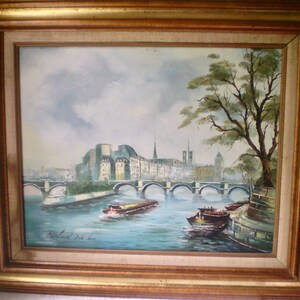 May include: An oil painting in a gold-toned wooden frame depicting a river scene with a multi-arched stone bridge, city buildings, and boats on the water. A large tree stands on the right bank. The artist's signature is visible in the bottom left.