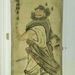 May include: An antique scroll painting depicting a figure in traditional garb, holding a sword. The figure has a beard and is wearing a hat with feathers. The painting is in muted tones of beige, black, and white, with Japanese text.