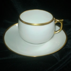 May include: White teacup and saucer set with gold trim. The cup has a rounded shape with a gold handle. The saucer is round and also has gold trim. The set is likely made of porcelain.