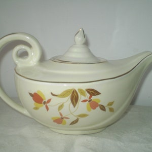 May include: A white teapot with a gold trim and a floral design featuring brown, orange, and yellow leaves. The teapot has a unique curved spout and a handle that loops back on itself.