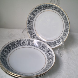 May include: A set of white porcelain plates with a black floral border and gold trim. The plates are stacked and displayed on a clear stand, showcasing their elegant design. The plates are suitable for serving appetizers or desserts.