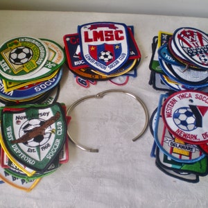 May include: A selection of soccer club patches in various colours, shapes, and designs. The patches feature team names, logos, and the text "SOCCER CLUB". A silver metal ring is also present.