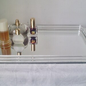 May include: A rectangular mirrored tray with a clear, ridged border. Several perfume bottles and a bottle of nail polish with gold caps are arranged on the tray. The tray is on a white surface.