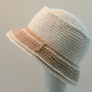 May include: A crocheted bucket hat with a wide brim. The hat is made with two shades of yarn, a light beige and a darker beige. The hat is tilted to the side.