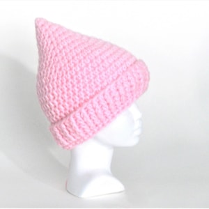 Może przedstawiać: Różowa czapka z dzianiny z ostrym wierzchołkiem. Czapka znajduje się na białej głowie manekina.