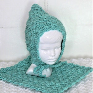 May include: A teal blue crocheted baby bonnet with a matching scarf. The bonnet has a pointed top and the scarf is tied at the neck.